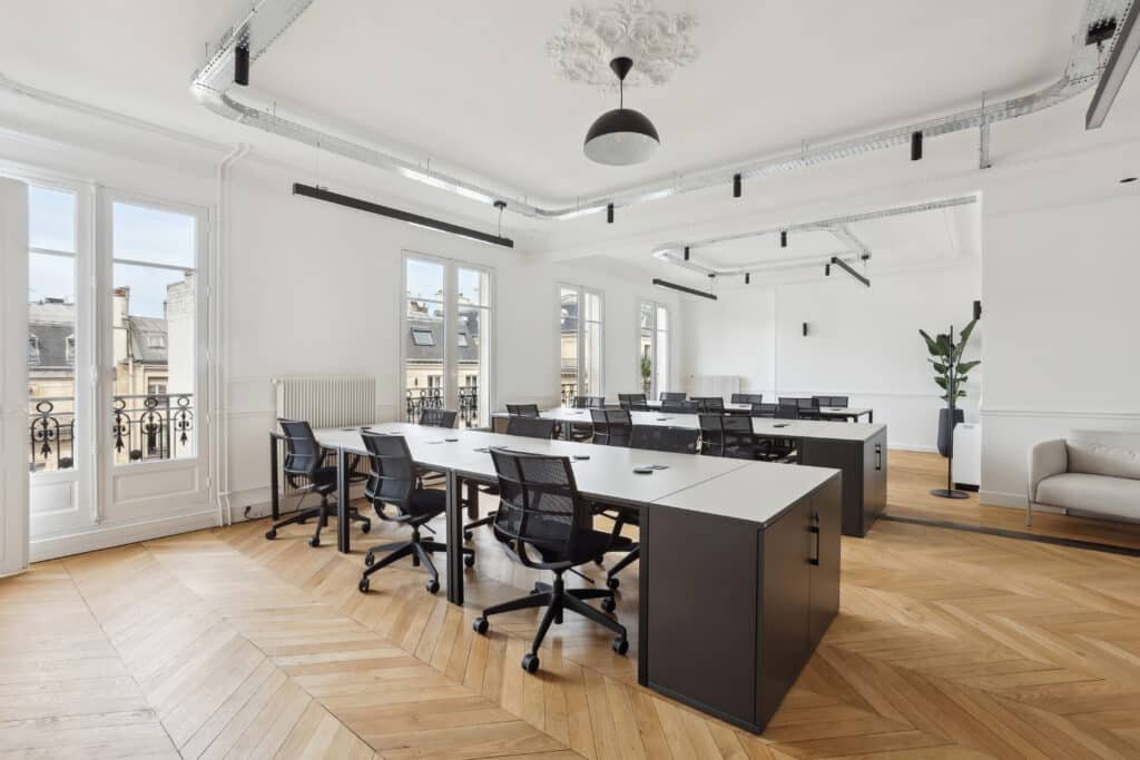 Un espace de bureau lumineux avec de grandes fenêtres, des bureaux alignés et des chaises noires, situé dans un bâtiment parisien avec vue sur les toits environnants.