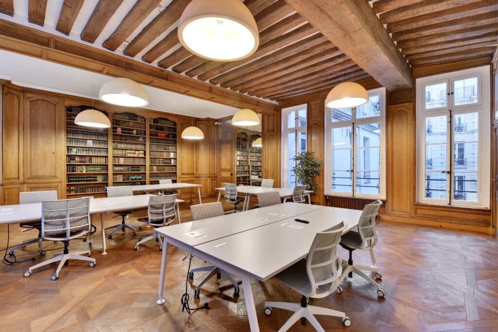 Salle de travail spacieuse avec des bureaux blancs, des chaises ergonomiques et des étagères en bois remplies de livres, située dans une pièce avec des plafonds en bois et un sol en parquet.