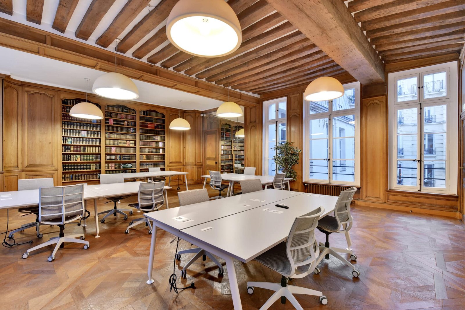 Salle de travail spacieuse avec des bureaux blancs, des chaises ergonomiques et des étagères en bois remplies de livres, située dans une pièce avec des plafonds en bois et un sol en parquet.